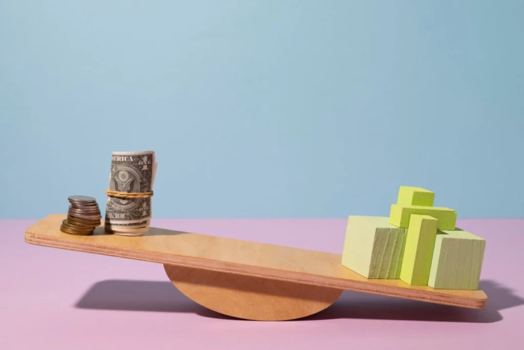 A stack of coins on a wooden seesaw on a pink background.