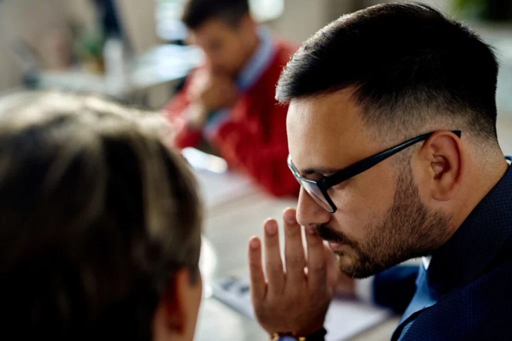 A man in glasses is talking to another man in a meeting.