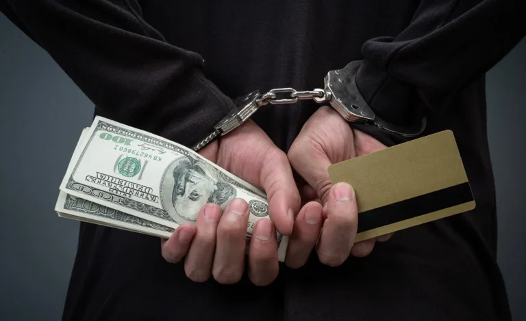 Person handcuffed holding a stack of U.S. dollar bills and a blank credit card.