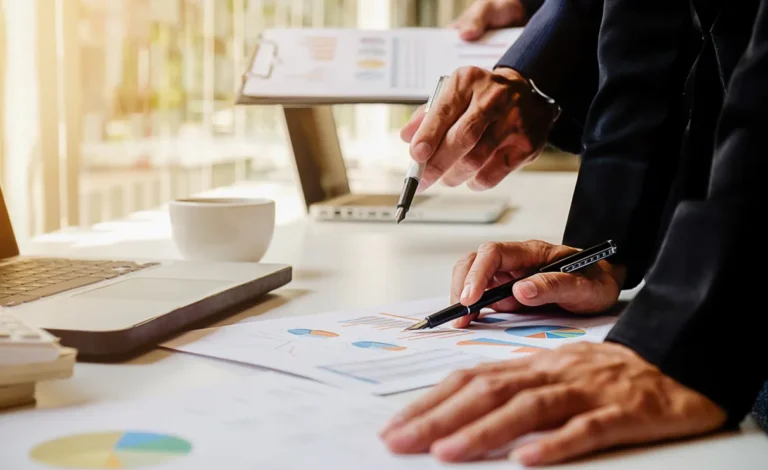 Two people analyzing charts and graphs with pens on a table, surrounded by a laptop and a cup, delve into financial services data.