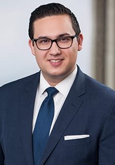 A man with dark hair and glasses wearing a dark blue suit, white shirt, and blue tie stands in front of a light background.