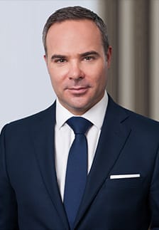 A man in a dark suit, white shirt, and blue tie stands in front of a neutral background, looking at the camera.