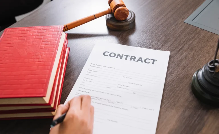A hand signing a contract document on a wooden desk, possibly addressing wrongful dismissal, with a stack of red books and a gavel nearby.