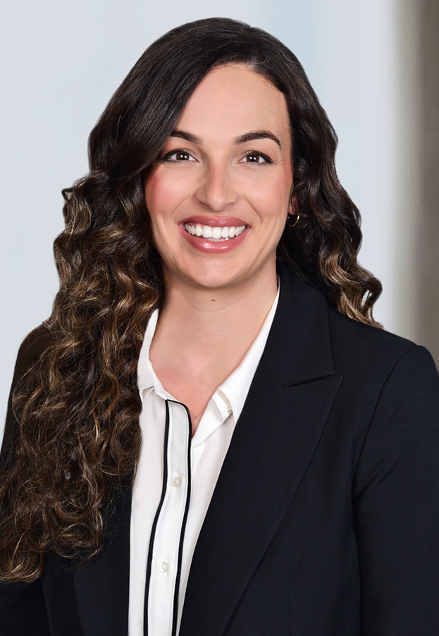 A person with long curly hair wearing a black blazer and a white shirt smiles against a plain background.