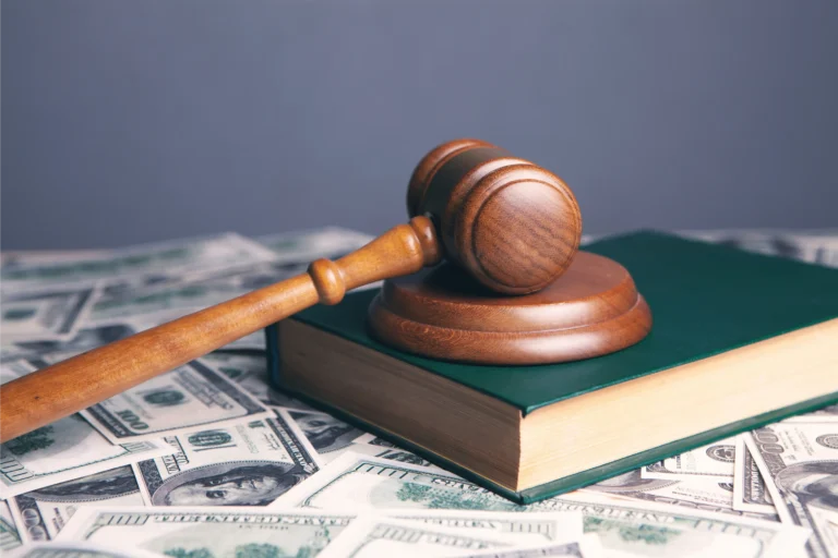 A wooden judge’s gavel rests on a closed book placed on top of scattered U.S. hundred-dollar bills.